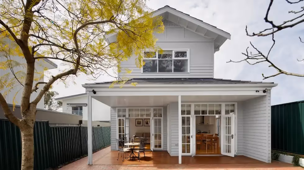 Two-storey home extension with light grey weatherboard cladding, covered patio, and French doors opening to an outdoor dining area.