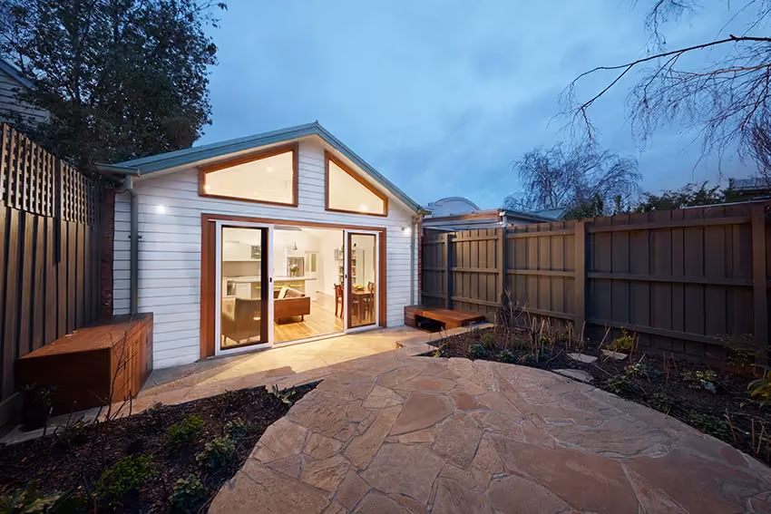 A small modern backyard extension with white horizontal cladding and large glass sliding doors glowing warmly at dusk. A paved stone patio and garden beds sit within a fenced courtyard under a blue evening sky.