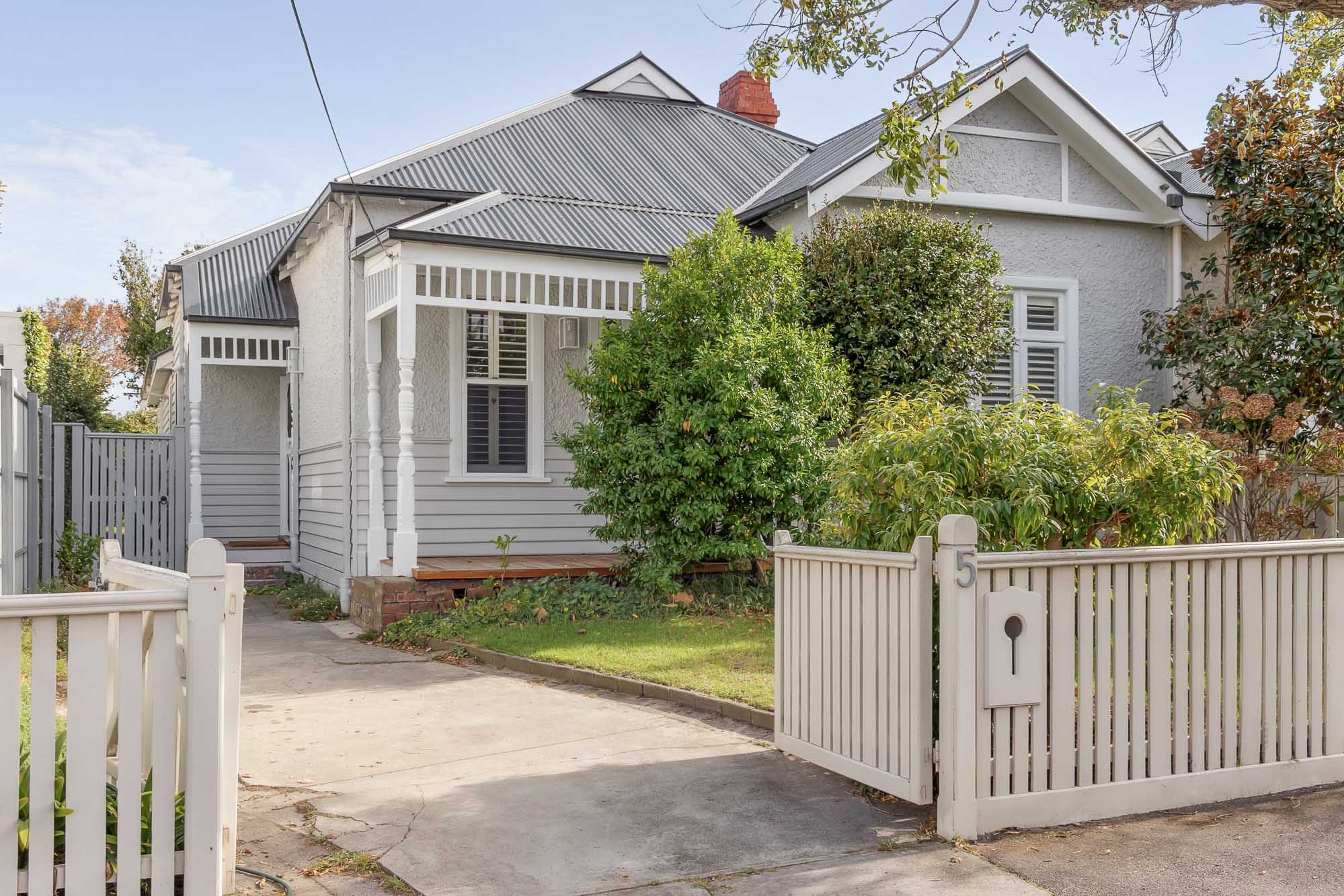 A single-storey light grey weatherboard house with a corrugated metal roof and small front veranda. A white picket fence and gate open to a concrete driveway, with green shrubs and trees in the front garden.