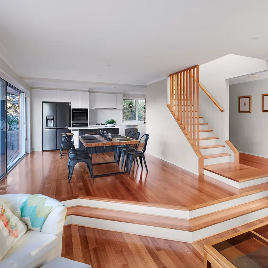 Modern open-plan kitchen and dining area with timber floors, white cabinetry, island bench, and a wooden staircase leading upstairs.