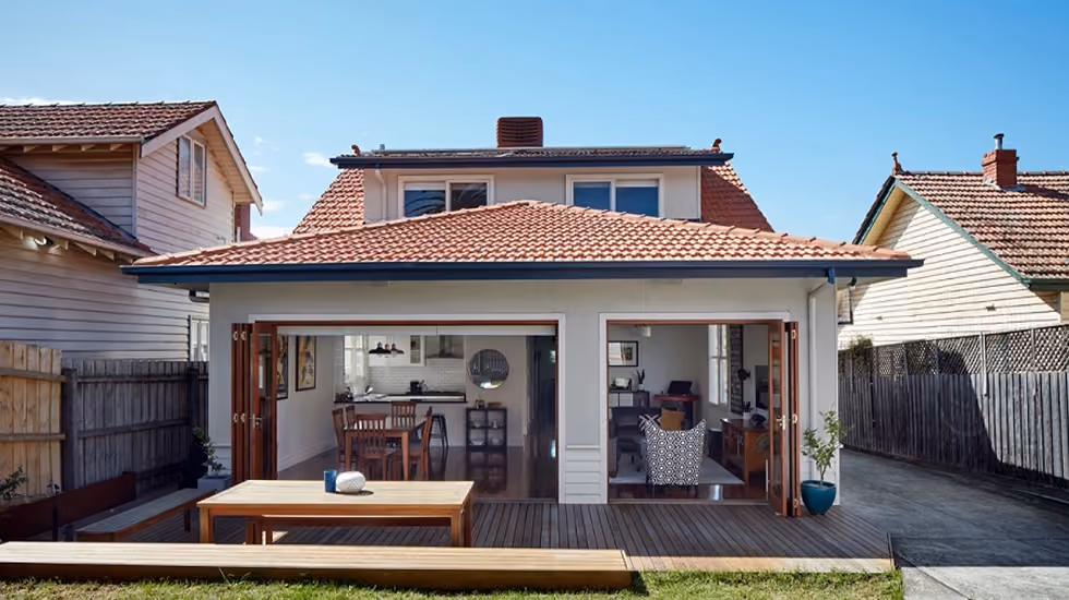 A rear exterior of an Australian house with a deck, yard, and bi-fold doors open to an indoor kitchen and living area.