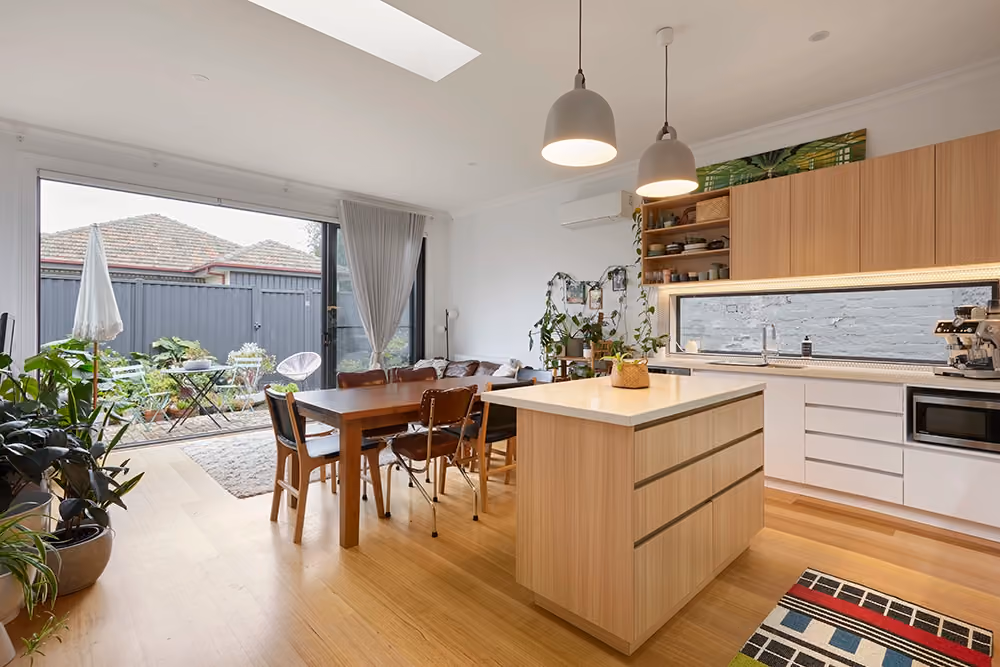 A bright, modern kitchen and dining area with light wood flooring, an island bench, and sliding doors opening to a small backyard garden.