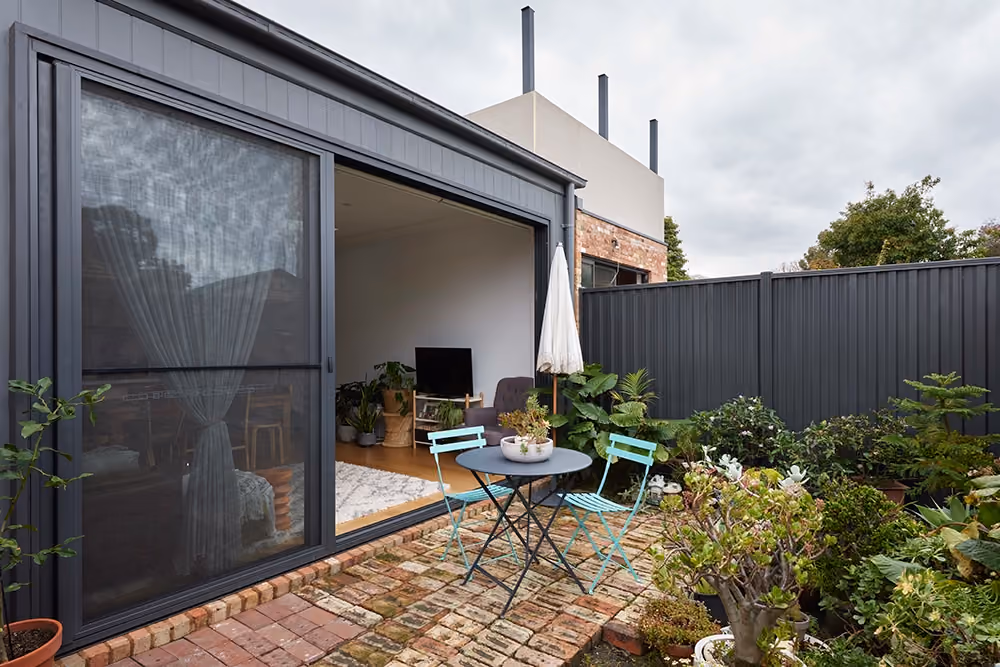 An exterior view of a modern house opening onto a small brick patio with aqua bistro chairs, surrounded by lush potted plants.