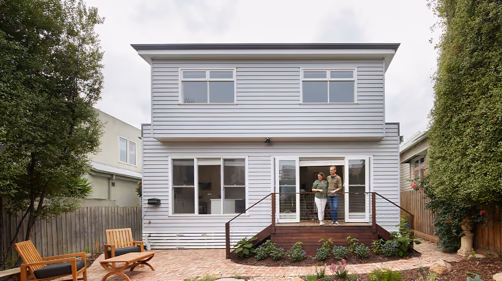 A two-story grey weatherboard house in an Aussie backyard, with a couple standing on the timber deck overlooking a small pond and paved area.