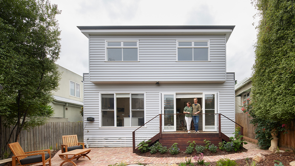 A two-story grey weatherboard house in an Aussie backyard, with a couple standing on the timber deck overlooking a small pond and paved area.