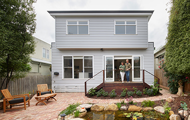 A two-story grey weatherboard house in an Aussie backyard, with a couple standing on the timber deck overlooking a small pond and paved area.