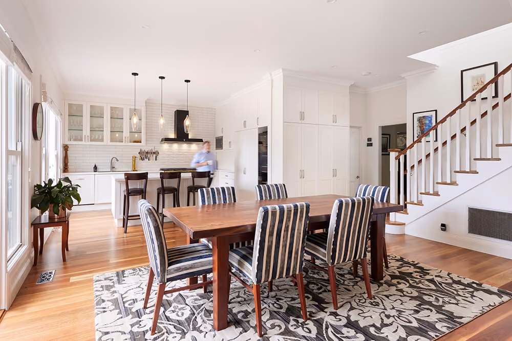 A bright, open-plan dining and kitchen area with polished timber floors. A wooden table with striped chairs sits on a patterned rug.