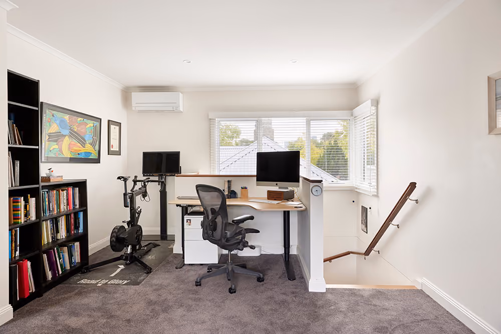 An elevated home office with a black standing desk, ergonomic chair, multiple screens, and a stationary exercise bike on carpet.