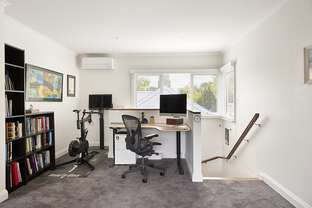 An elevated home office with a black standing desk, ergonomic chair, multiple screens, and a stationary exercise bike on carpet.