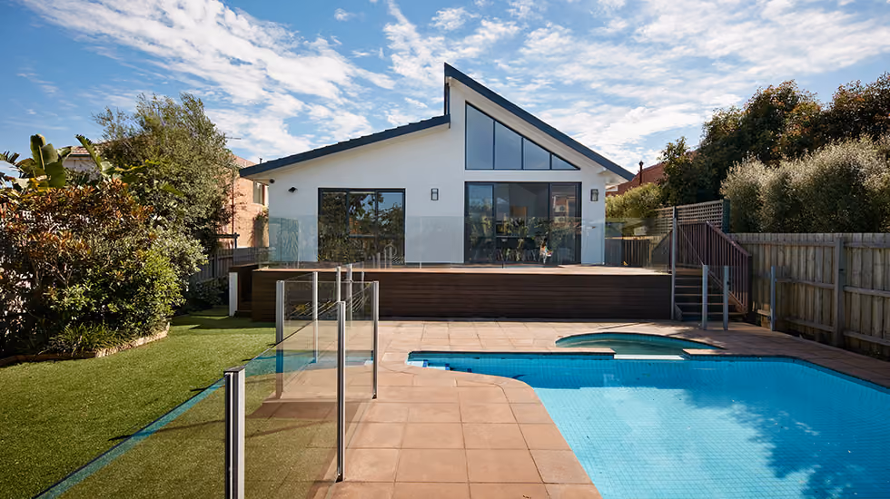 A modern white house with a unique roofline and large glass doors opening onto a wooden deck overlooking a swimming pool and a lush green lawn.