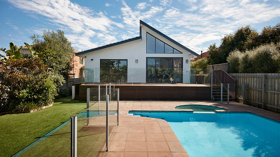 A modern white house with a unique roofline and large glass doors opening onto a wooden deck overlooking a swimming pool and a lush green lawn.