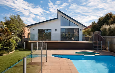 A modern white house with a unique roofline and large glass doors opening onto a wooden deck overlooking a swimming pool and a lush green lawn.