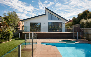 A modern white house with a unique roofline and large glass doors opening onto a wooden deck overlooking a swimming pool and a lush green lawn.
