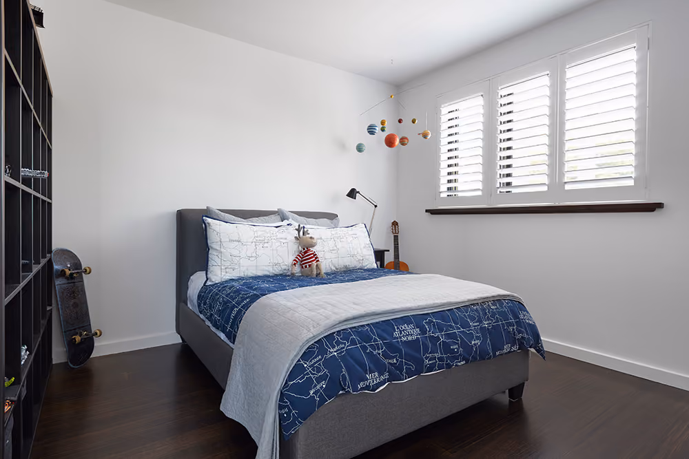A bright, modern boy's bedroom with a dark blue and white map-themed bedspread, white window shutters, and a solar system mobile.