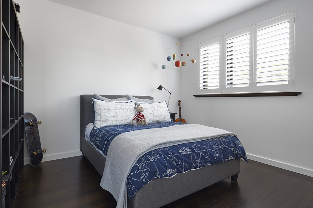 A bright, modern boy's bedroom with a dark blue and white map-themed bedspread, white window shutters, and a solar system mobile.