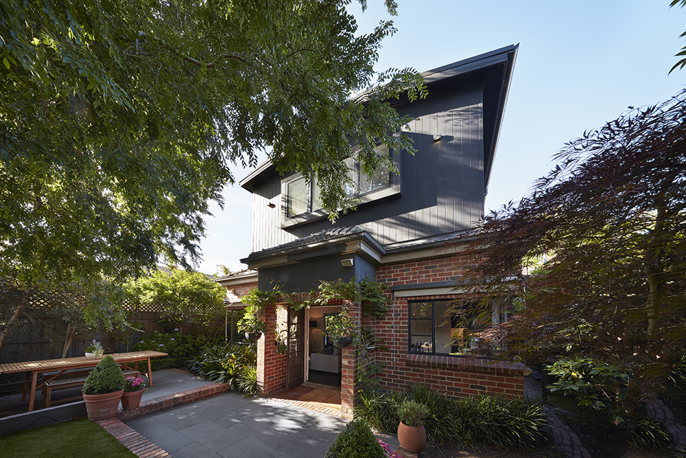Two-storey home with dark cladding and red brickwork. Patio, wooden table, and lush greenery surround the entrance.