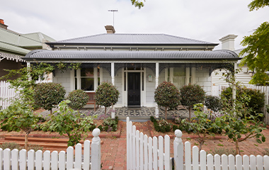 A modern white house with a unique roofline and large glass doors opening onto a wooden deck overlooking a swimming pool and a lush green lawn.