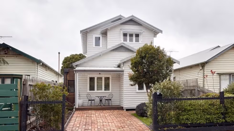 A 2-storey light grey weatherboard home featuring a paved brick pathway, dark fence, and an outdoor table setting.