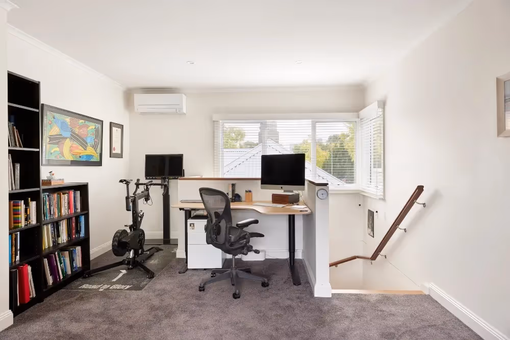 Upper-floor home office featuring a computer desk, ergonomic chair, stationary bike, black bookcases, and carpeted floor.