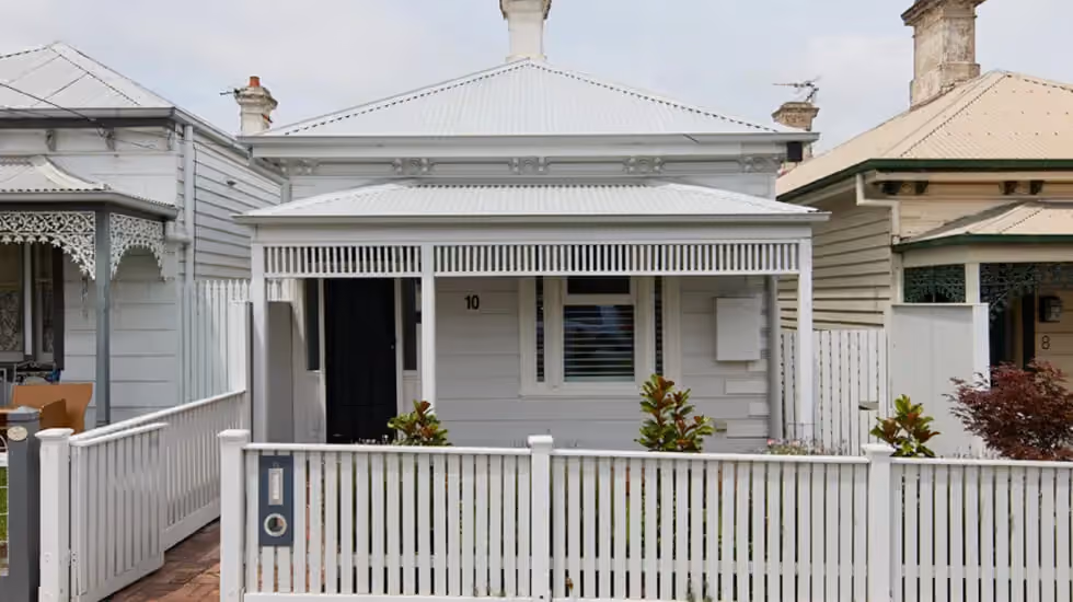 A single-storey white weatherboard cottage with a metal roof, front veranda, and picket fence on a suburban street.
