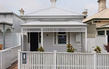 A single-storey white weatherboard cottage with a metal roof, front veranda, and picket fence on a suburban street.