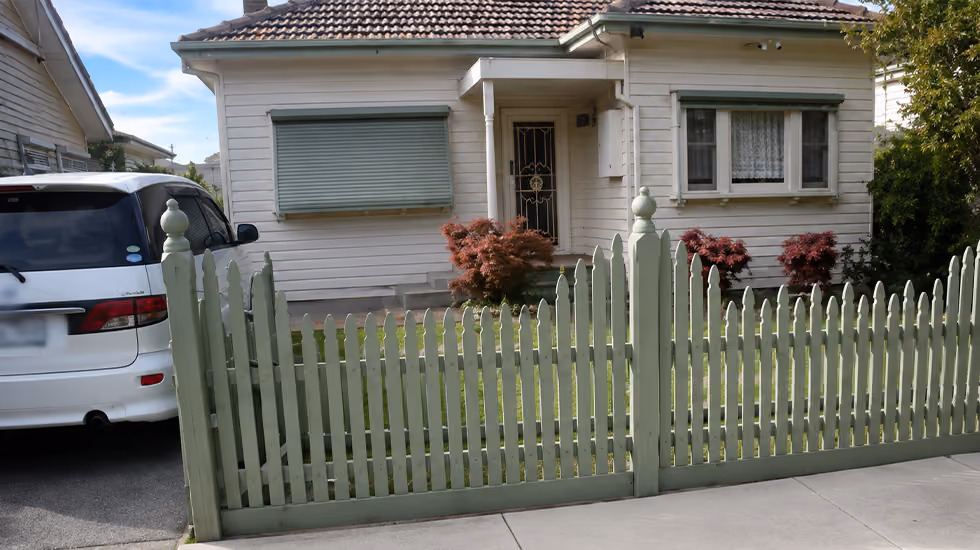 A single-storey white weatherboard house with a tiled roof, front window shutters, and a green timber picket fence.