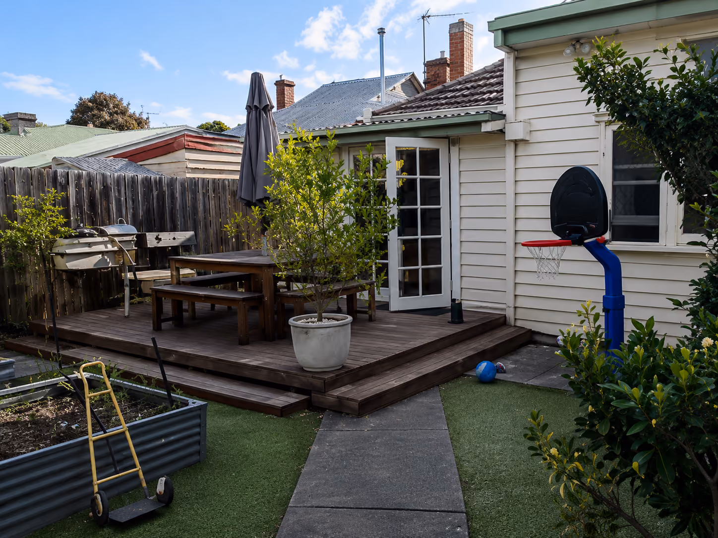 A backyard with a wooden deck, outdoor dining setting, glass French doors, and a small basketball hoop near the lawn.