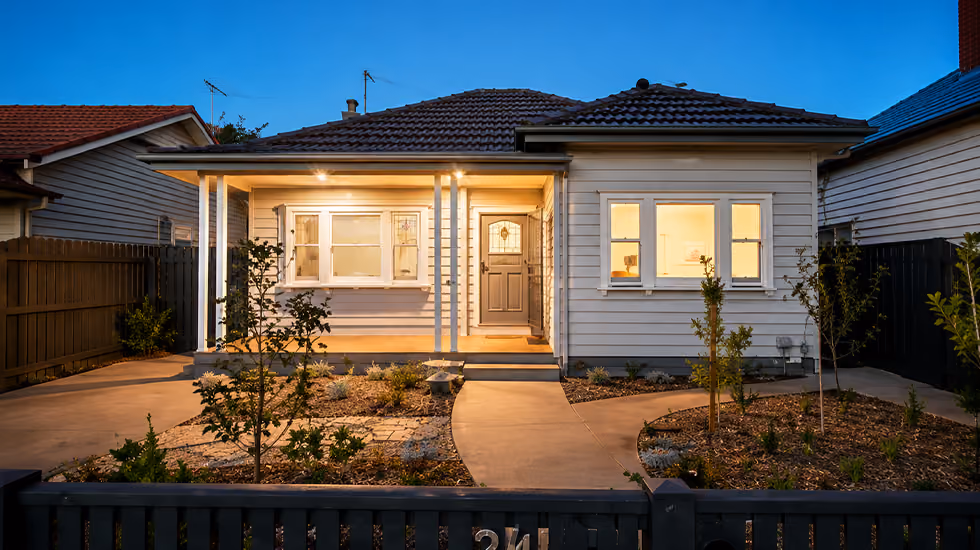 A renovated single-storey weatherboard home lit up at dusk with a landscaped front yard and dark picket fence.