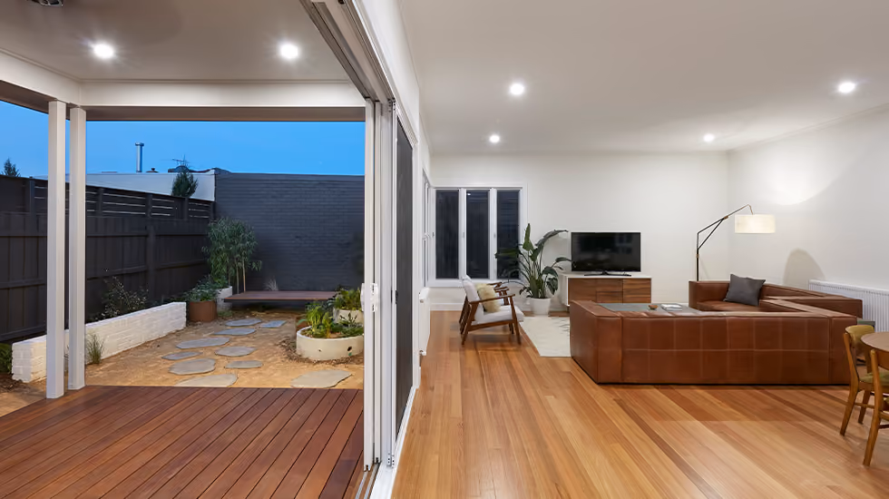 A modern open-plan living room with timber floors opening onto a covered deck and courtyard with stepping stones.