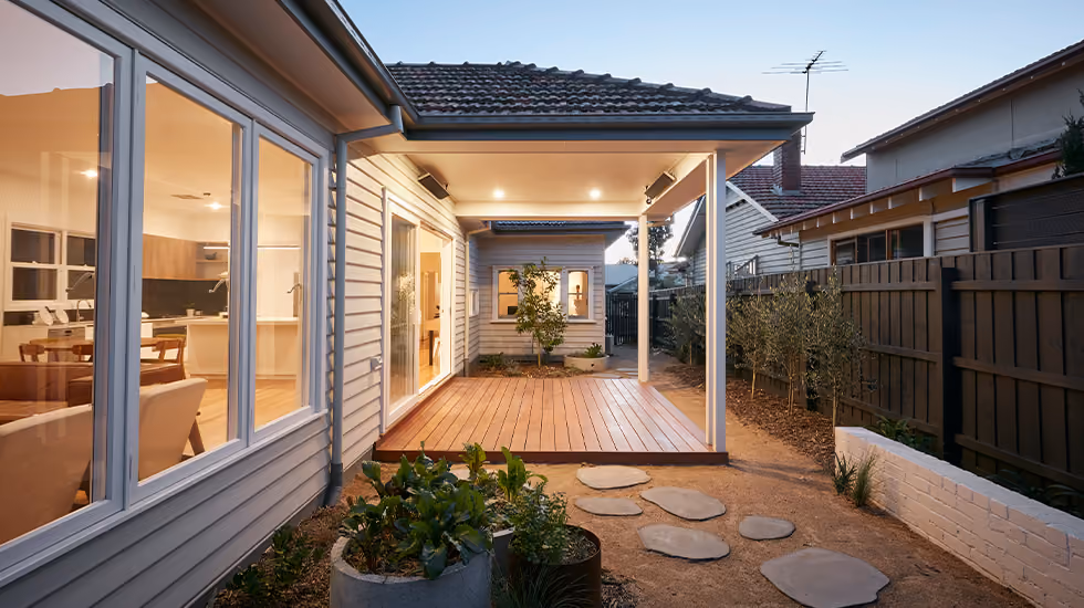 Exterior view of a weatherboard home showing large glass windows, a covered outdoor deck, and paved courtyard at dusk.]