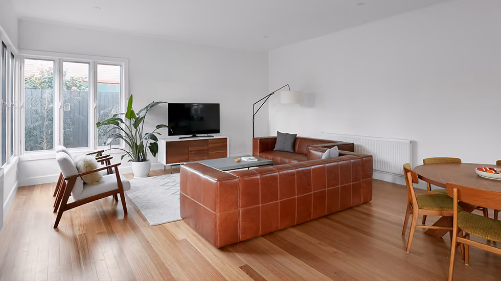 A modern living room featuring a tan leather modular sofa, white walls, timber floorboards, and a large indoor plant.