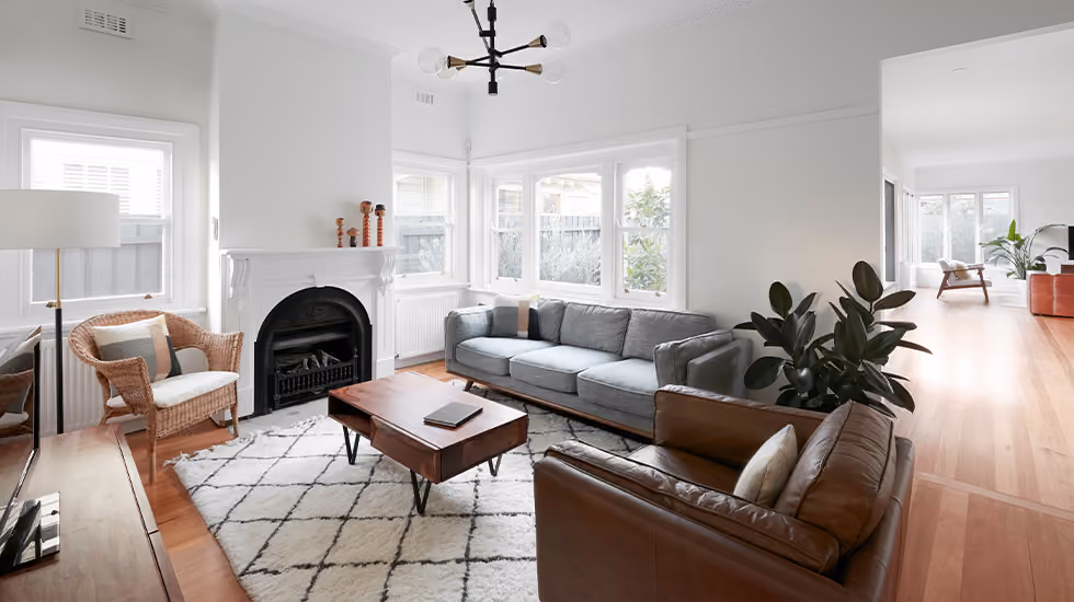 A stylish living room featuring a grey lounge, black period fireplace, patterned rug, timber flooring, and pendant light.]