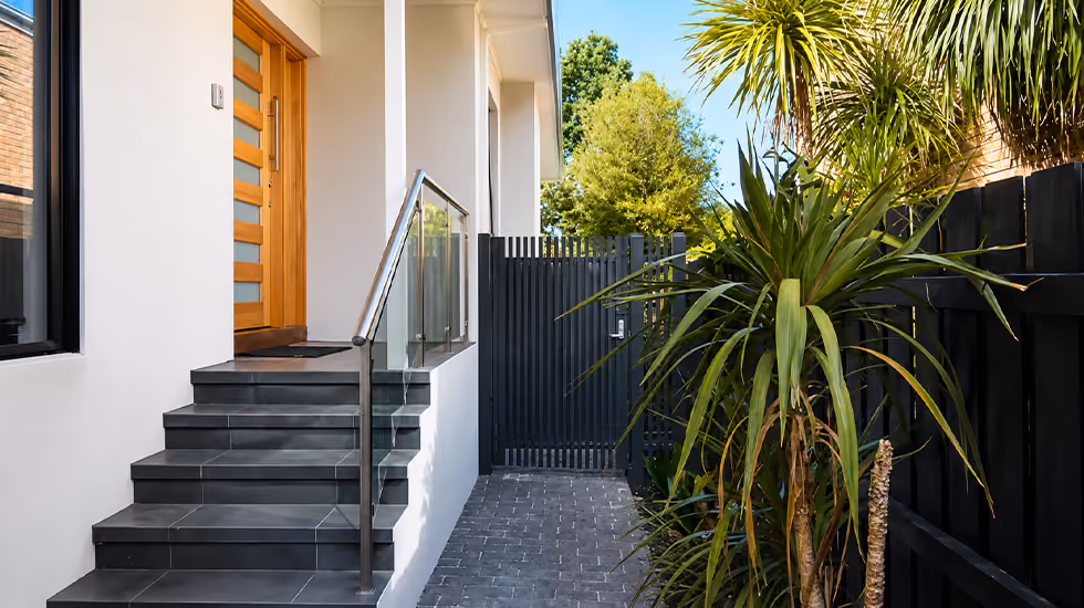 A modern home entrance with dark tiled steps, glass balustrade, wooden front door, and lush tropical plants by a fence.