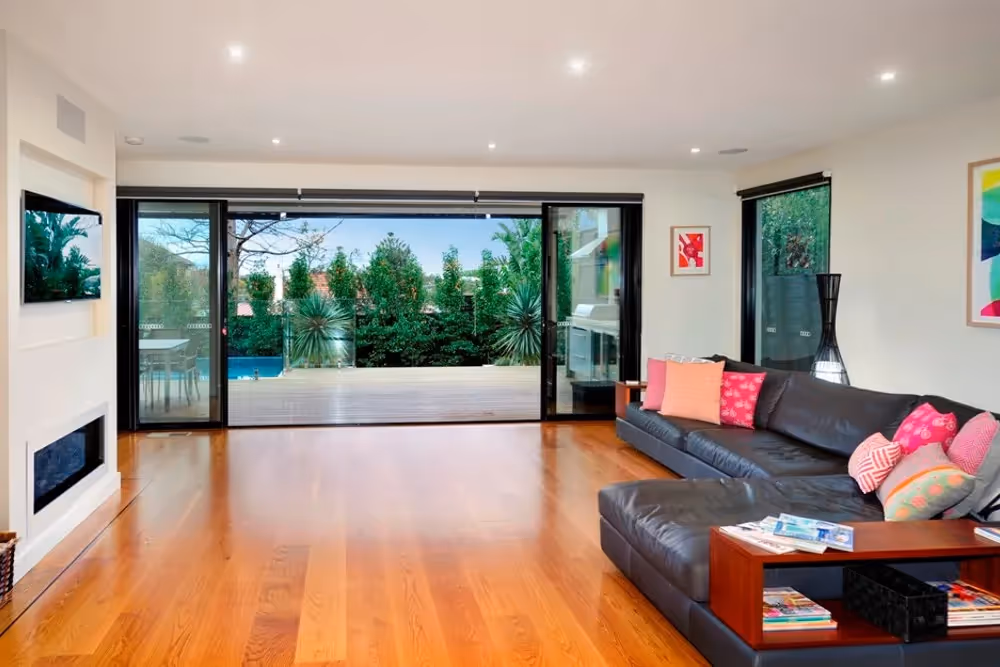 An open-plan living area with timber flooring, a dark L-shaped sofa, and sliding glass doors opening to an outdoor deck.