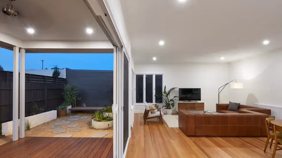 A modern open-plan living room with timber flooring opening onto a covered deck and courtyard with stepping stones at dusk.
