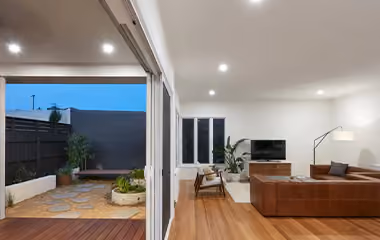 A modern open-plan living room with timber flooring opening onto a covered deck and courtyard with stepping stones at dusk.