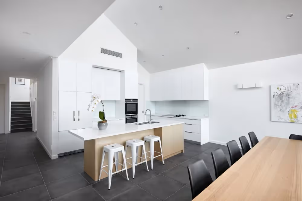 A contemporary white kitchen with high vaulted ceilings, a central island bench with bar stools, and dark tiled flooring.
