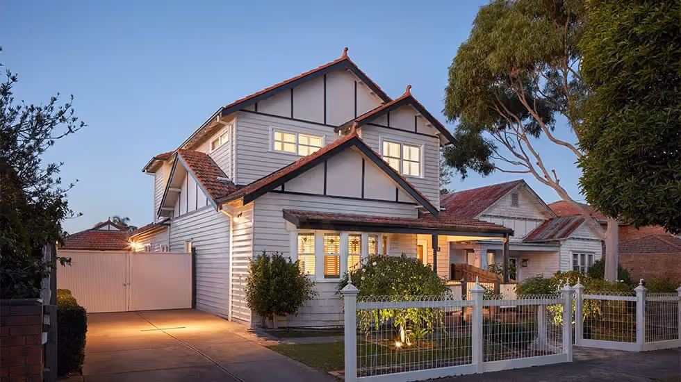 A double storey, white weatherboard house with terracotta roof tiles and an illuminated front yard behind a white fence at dusk.