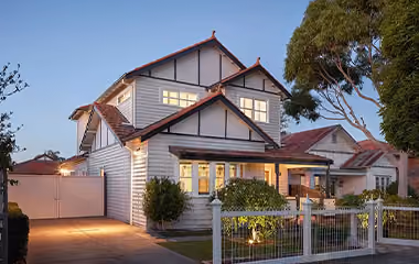 A double storey, white weatherboard house with terracotta roof tiles and an illuminated front yard behind a white fence at dusk.