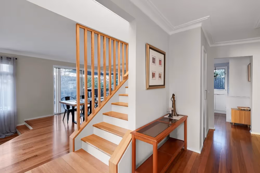 A modern hallway featuring a timber staircase with a vertical slat balustrade, polished wooden floors, and a console table.