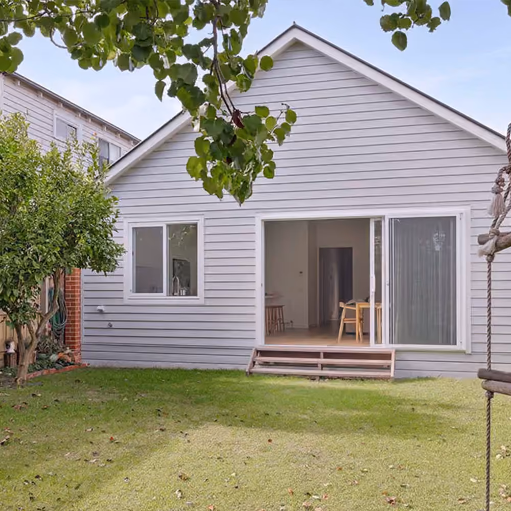Rear exterior of a ground floor home extension with grey weatherboard cladding, sliding glass doors and a backyard lawn.