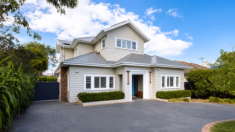 A two-storey light grey weatherboard home with a metal roof, plantation shutters, and a wide paved driveway on a clear day.
