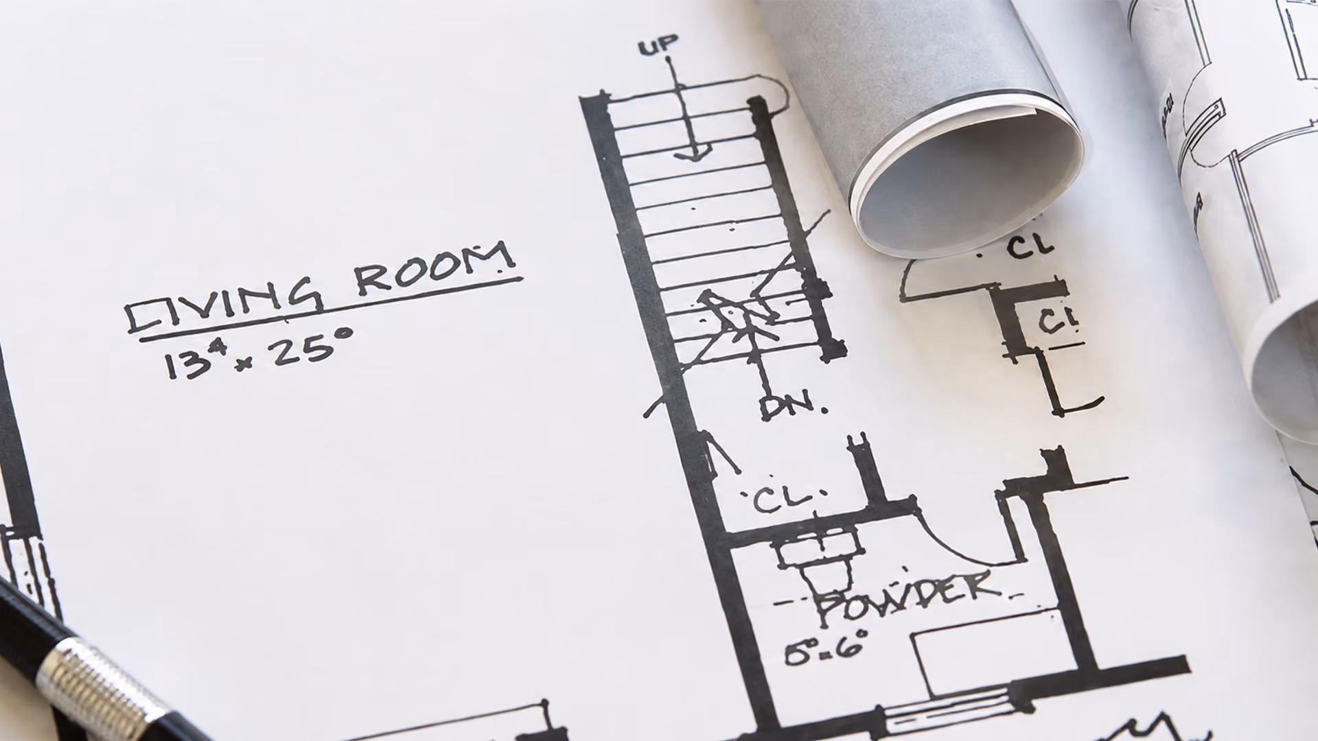Hand-drawn architectural floor plan sketch showing a living room layout with rolled-up blueprints and a mechanical pencil.