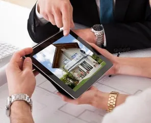 Two people in business attire view a house image on a tablet, with one person pointing at the screen during a property discussion at a desk.