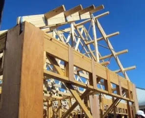 Timber house frame under construction with exposed wooden studs and roof trusses against a clear blue sky.