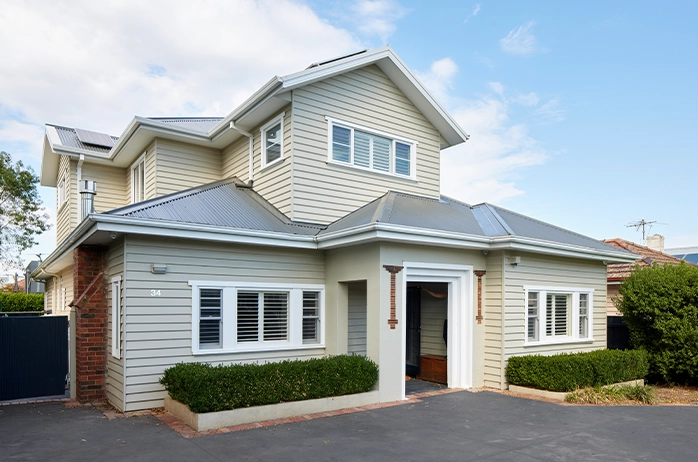 Exterior view of a light grey weatherboard house with a metal roof, white-framed windows with shutters, and a recessed front entry, set behind low hedges and a paved driveway under a partly cloudy sky.