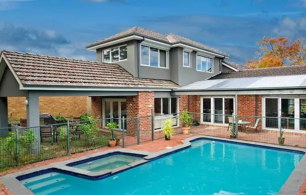 Exterior view of a light grey weatherboard house with a metal roof, white-framed windows with shutters, and a recessed front entry, set behind low hedges and a paved driveway under a partly cloudy sky.