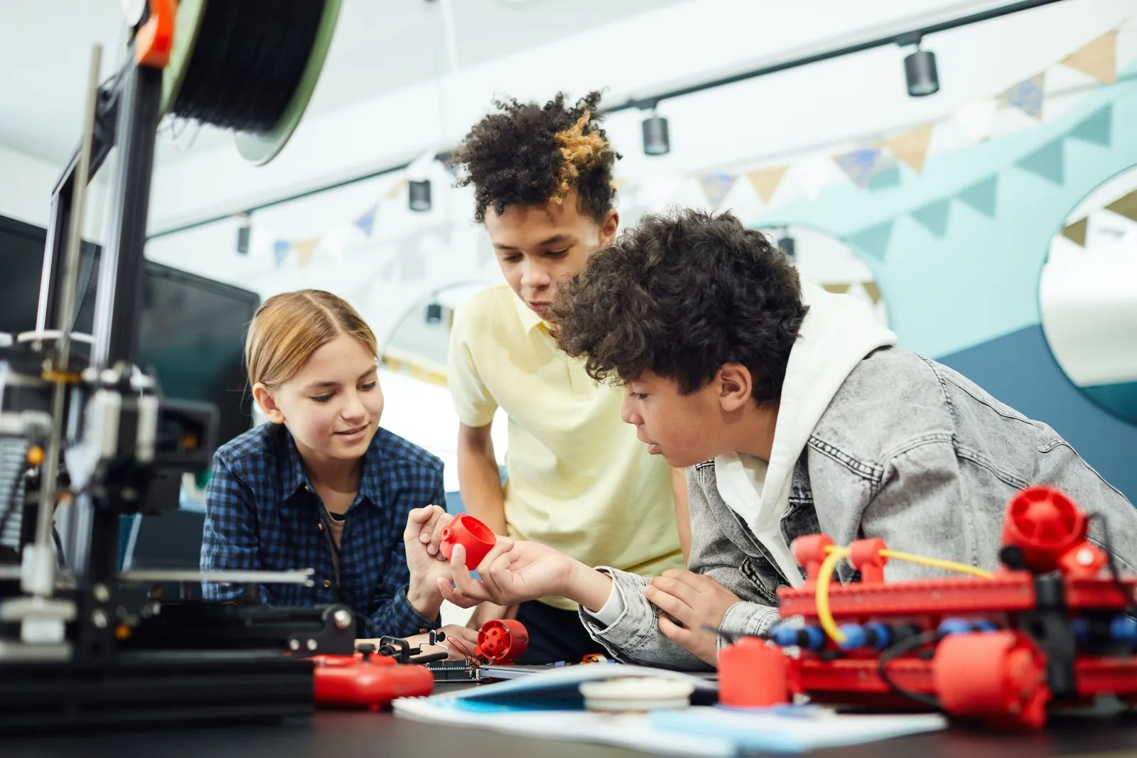 A 3D printed part being examined at a workbench alongside other printed pieces and a 3D printer