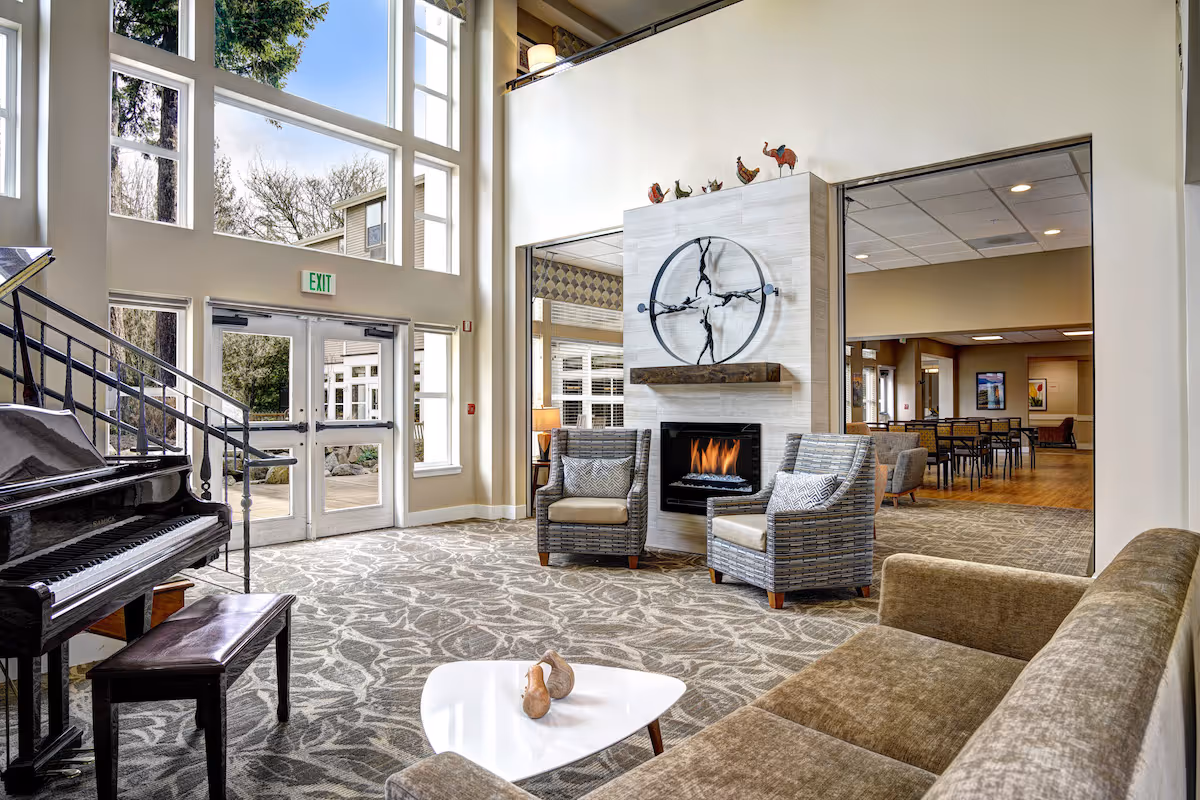 Studios in Federal Way, WA | Federal Way | Lobby with Carpeted Flooring, Cozy Seating, Piano, Fireplace, Large Windows, and Stylish Decor