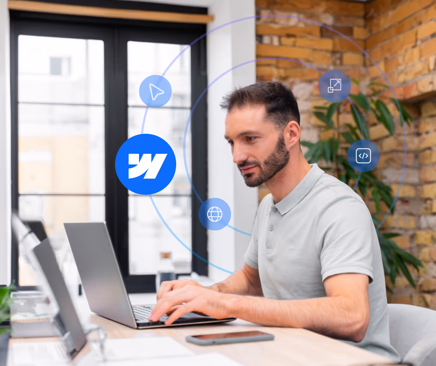 Man in gray polo shirt typing on a laptop at a wooden desk in a bright office with brick walls and digital icons floating around him.
